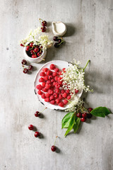 Fresh cherry and raspberry berries in ceramic mug and plate, elderflowers, jug of cream over gray texture background. Flat lay, space