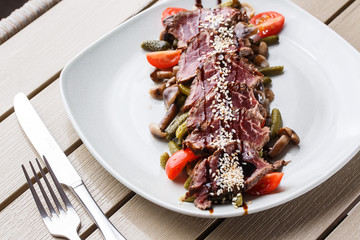 Sliced medium rare Roast beef with pickles, cherry tomatoes,mushrooms on a white plate on wooden background.