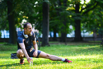 Fototapeta premium Fit man doing stretching exercises outdoors, young male sportsman stretching and preparing to run, attractive adult runner stretching the legs standing on grass in the park, sports fitness concept