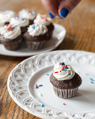 Chocolate mini cupcake on white plate being decorated with red, white, and blue sprinkles.  Fingers with motion blurr in photo.  Additional decorated cupcakes blurred in background.