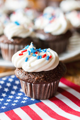 Close up of chocolate mini cupcake decorated with red, white, and blue sprinkles.  US flag featured.  Additional cupcakes blurred in the background.  Wooden table.