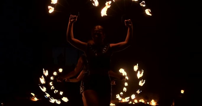 Three women with burning hoops dance with fiery torches in leather clothes in a dark hangar demonstrating a circus fire show in slow motion.