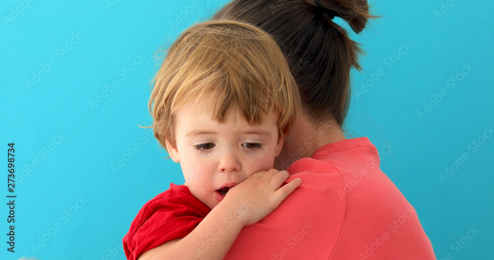 Wall mural Side view of young mom cuddling sad kid with finger in mouth looking down isolated on blue background