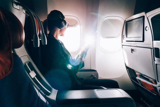 Side view of businesswoman in elegant wear looking in airplane window and waiting take-off from airport during trip to conference, intelligent female in electronic headphones listening audio book