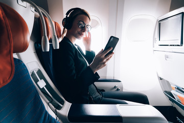 25 years old young woman traveler making WiFi purchase on electronic device in airplane, female passenger holding mobile phone in aircraft and checking wifi connection while listening audio book
