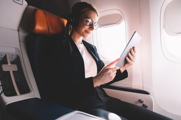 Positive young woman sitting in passenger seat in airplane and reading interesting book via...