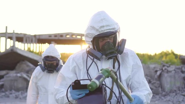 scientists in protective suits and masks and a dosimeter , walk measure radiation on the background of the ruins of a man-made disaster close up at sunset