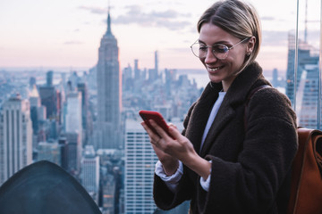Smiling caucasian female using application in roaming on mobile phone while standing on Observation deck viewpoint with Empire State view building during vacations.Happy woman tourist in New York city - obrazy, fototapety, plakaty