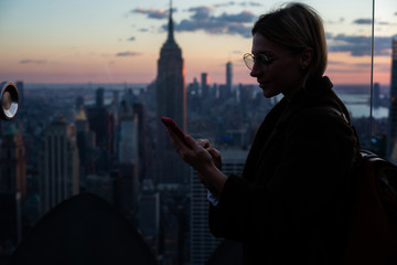 Hipster girl in autumn coat using smartphone roaming network while standing on Observation deck...