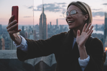 Happy caucasian hipster girl saying Hello to her followers in travel blog while visiting famous landmark place with scenery view of New York cityscape. Young woman tourist enjoying journey to USA