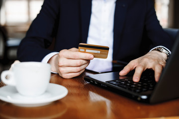 Male hand holding a credit card while operating at a laptop sitting at a desk drinking coffee.