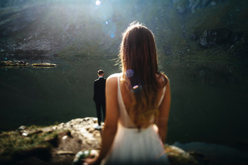 Back view of a unrecognizable groom sitting at the edge of a lake in the mountains while his bride is looking at him.