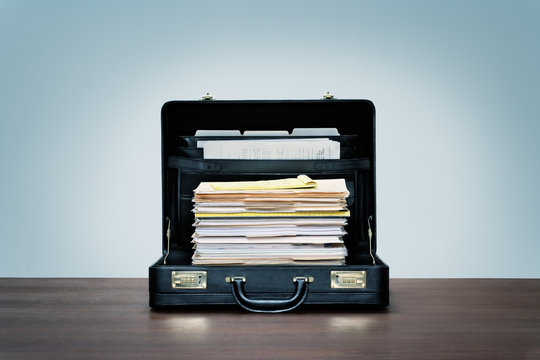 Tall stack of files and folders in black leather briefcase on wood desk with blue background.