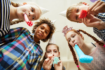 Low angle view at multi ethnic group of children blowing party horns at camera and having fun