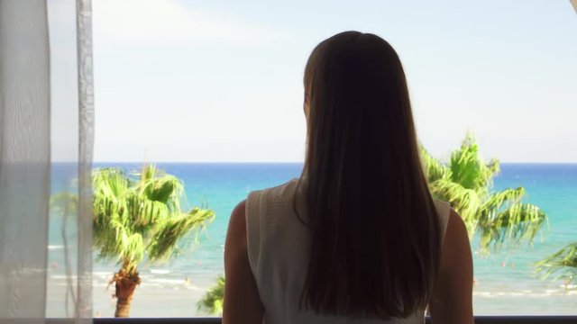 Young woman going out onto balcony and enjoying view from terrace. Female on vacation opening curtains and looking at Mediterranean sea. Wind swaying leaves on palm trees. Camera follows.