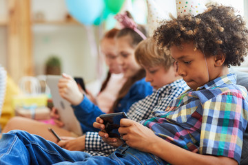 Obraz premium Side view at group of children using smartphones sitting in row at birthday party, copy space