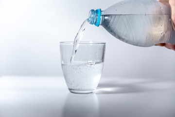 Hand pouring sparkling water from plastic bottle to glass filled with cold mineral water on bright, blank surface. Reflection of the glass visible on the surface. High exposure photo. Side white light