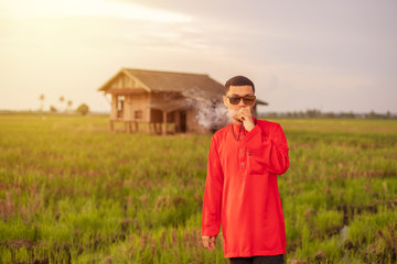 Portrait of young man smoking vape or vapor with background of traditional house in the middle of paddy field meadow.