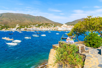 Couple of people sitting on coastal promenade in Cadaques village with port, Costa Brava, Spain