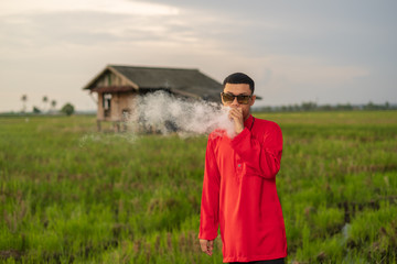 Portrait of young man smoking vape or vapor with background of traditional house in the middle of paddy field meadow.