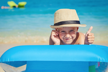 European boy in a sun hat is relaxing at the seashore, he is laying on the inflatable blue mattress, enjoying his summer holidays and showing like gesture.