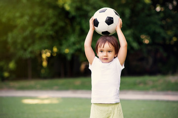 Little soccer player standing at football field outdoors: toddler boy holding ball above head ready...