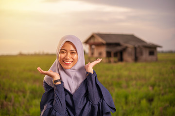 Close up portrait of happy young pretty Asian woman in hijab with paddy field meadow background and traditional house.Standing and smile. Copy space.