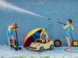 Funny children in boys' bathing suits posing in front of camera with high-pressure car wash and toy yellow car and scooters. Brothers are trying to wash car. Rainbow umbrella