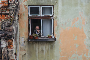 A senior man smokes in a wooden window of an old high-rise building with peeling walls. concept of past years. aging house, aging man with gray hair