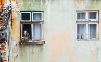 A senior man smokes in a wooden window of an old high-rise building with peeling walls. concept of past years. aging house, aging man with gray hair
