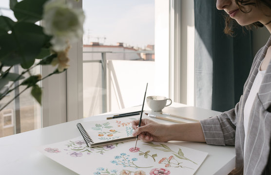 Crop person hand with brush painting watercolor flowers on large sheet at desk