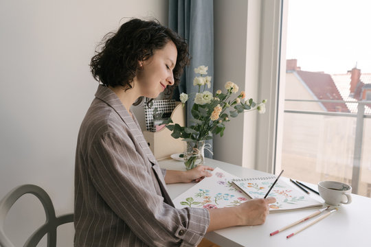 Elegant woman with brush painting watercolor flowers on large sheet at desk
