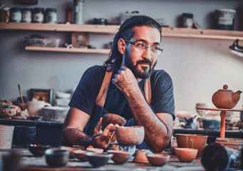 Diligent pensive man in glasses at his pottery workshop is workig for new project.