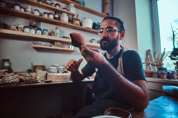Pensive entusiastici potter in glasses at his own workshop is workig on a new handmade teapot.