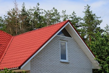 Fototapeta na wymiar gray brick attic with a window under a red tile roof against the sky and pine trees