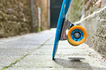 Detail of blue plastic mini cruiser penny board or skate board with yellow and orange wheels parked agains the wall in an alley. Low perspective, no people.   