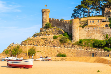 Fishing boats on golden sand beach in Tossa de Mar town with castle in background at sunset time,...