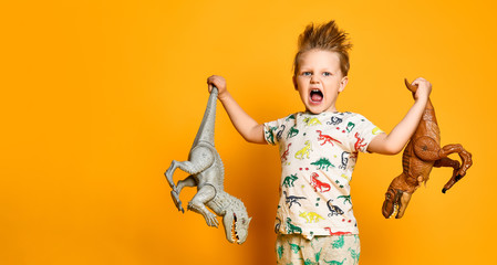 little cheerful boy holds a plastic dinosaur in each hand. The boy is dressed in a suit with...