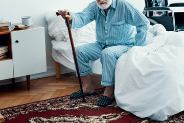 Old sick man with grey beard and hair wearing blue pajamas and sitting on bed at home