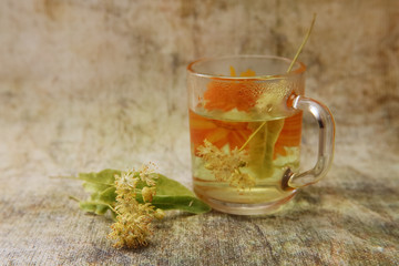 A cup of freshly linden tea. White vase with a bouquet of fragrant yellow flowers and leaves of linden and dried grass hedgehogs.