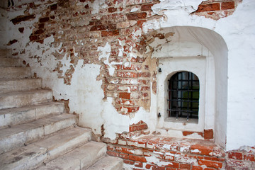 View of a part of stone steps in an old brick house. The brick wall is partially destroyed. Window opening with bars on the wall near the entrance to the house. Exfoliated white stucco on a brick wall