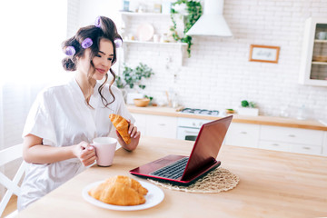 Fototapeta na wymiar Young carefree housewife sitting on table in kitchen. Having breakfast in morning. Holding cup of drink and croissant. Looking at laptop on table. Wear curlers in hair and white dressing gown.