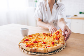 Cut view of woman in white dressing gown in kitchen. Taking piece of pizza. Small slice. Young housekeeper live carefree life.