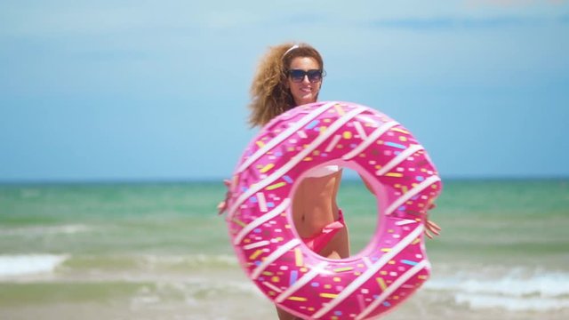 Woman having fun and smile dancing with pink donut. girl in bicini wearing sunglasses on the beach. Beautiful sexy woman on summer travel vacation.