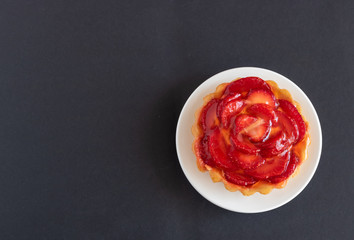 Tartalet with vanilla cream covered with slices of strawberry on the white plate on the black background. Top view.
