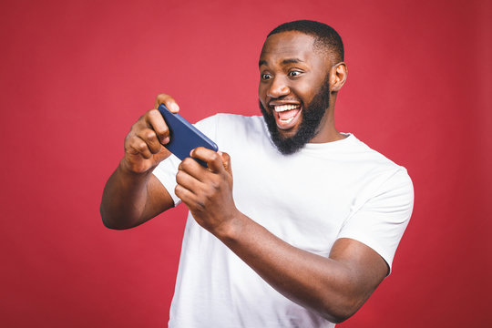 Portrait of a cheerful young african man dressed in casual playing games on mobile phone isolated over red background.
