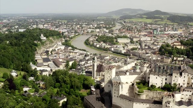 Flying over the Hohensalzburg in Salzburg, Austria to the old town and the river Salzach. The weather is sunny and people are walking through streets.