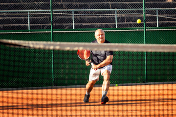 A middle-aged bald man emotionally plays tennis on the court. Outdoor.