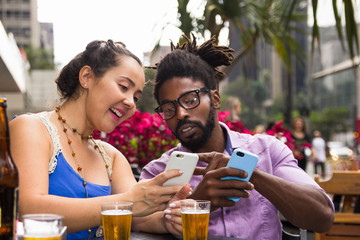 Multiracial couple at outdoor table in restaurant or bar drinking and using cell phone.