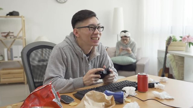 two college couple having fun themselves on summer break in dirty room. young man playing online video on computer using control while girlfriend relaxing on couch with mobile phone game in apartment